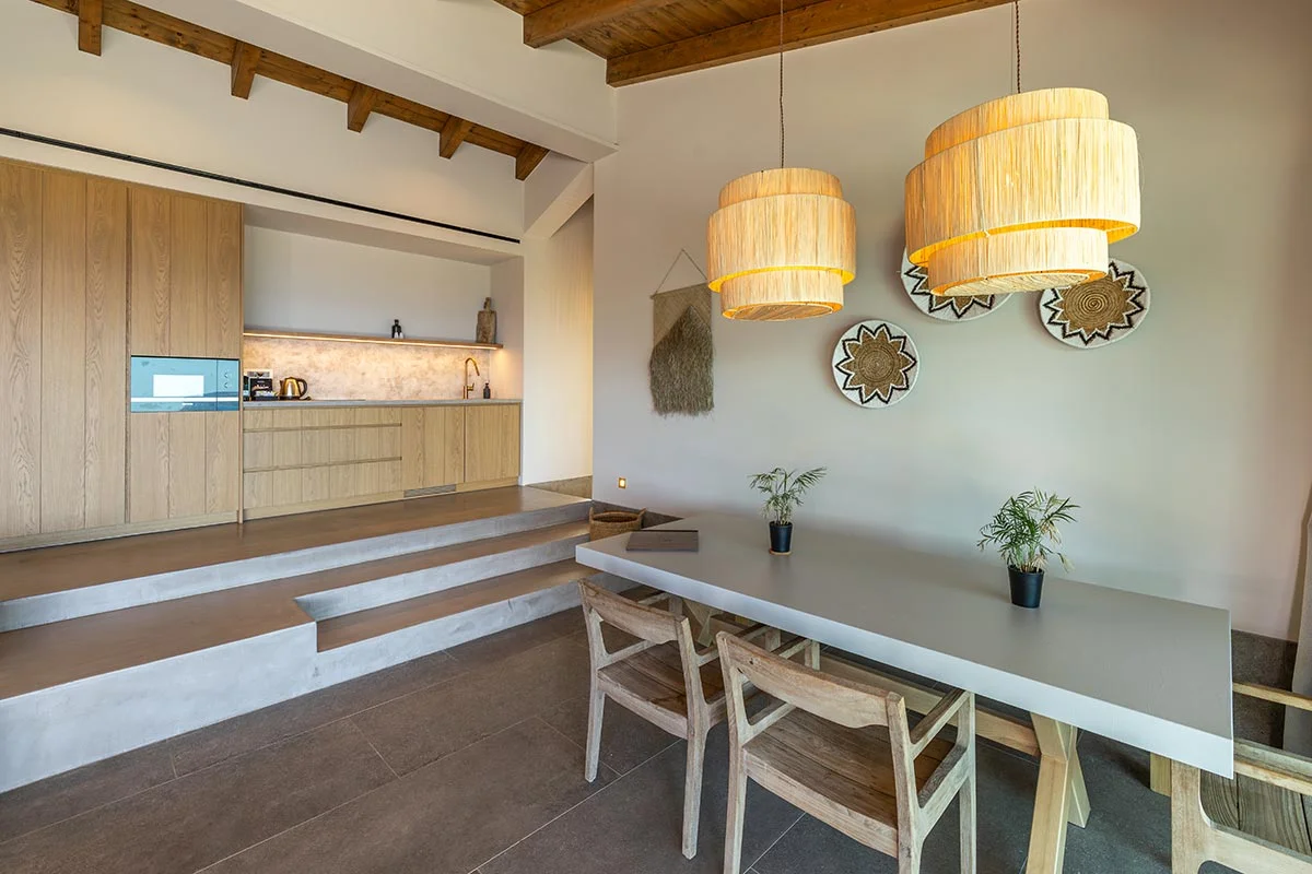 Dining area with wooden table, natural pendant lights, and open kitchen at Eliamos in Kefalonia, Greece.