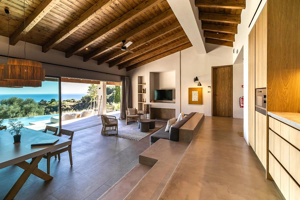 Open-plan villa interior at Eliamos Kefalonia with sea view, dining table, and exposed wooden ceiling beams.