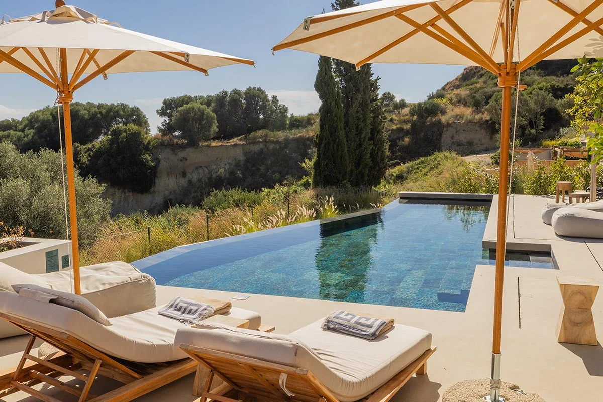 Private infinity pool with cushioned loungers and parasols overlooking natural hillside at Eliamos in Kefalonia.