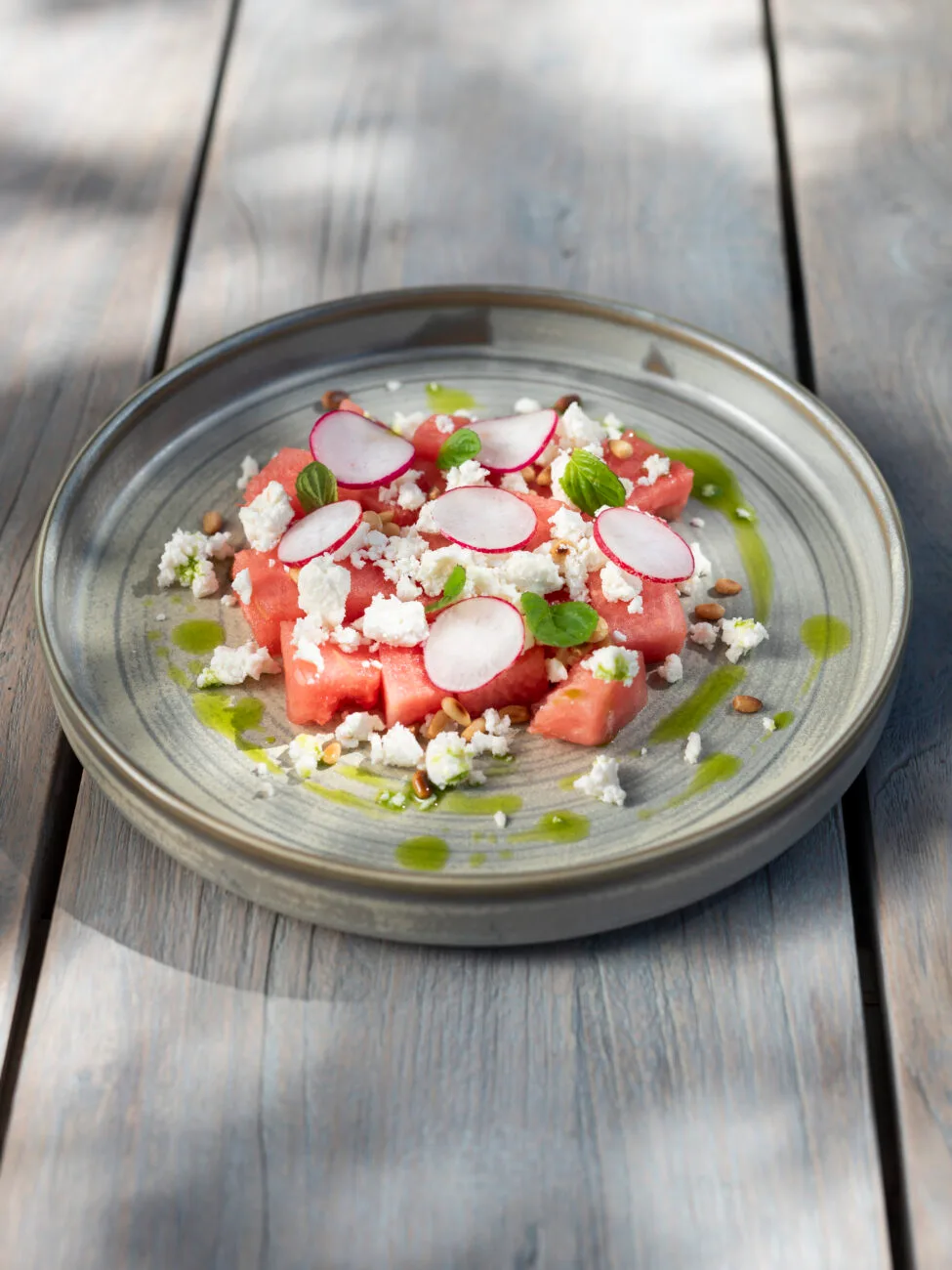 Watermelon salad with crumbled feta, sliced radishes, mint leaves, and pine nuts on a grey plate at Eliamos in Kefalonia.