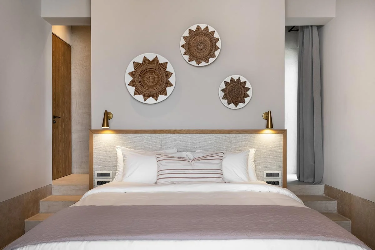 Bed with layered pillows and wall-mounted woven plates in a cosy neutral bedroom at Eliamos in Kefalonia, Greece.