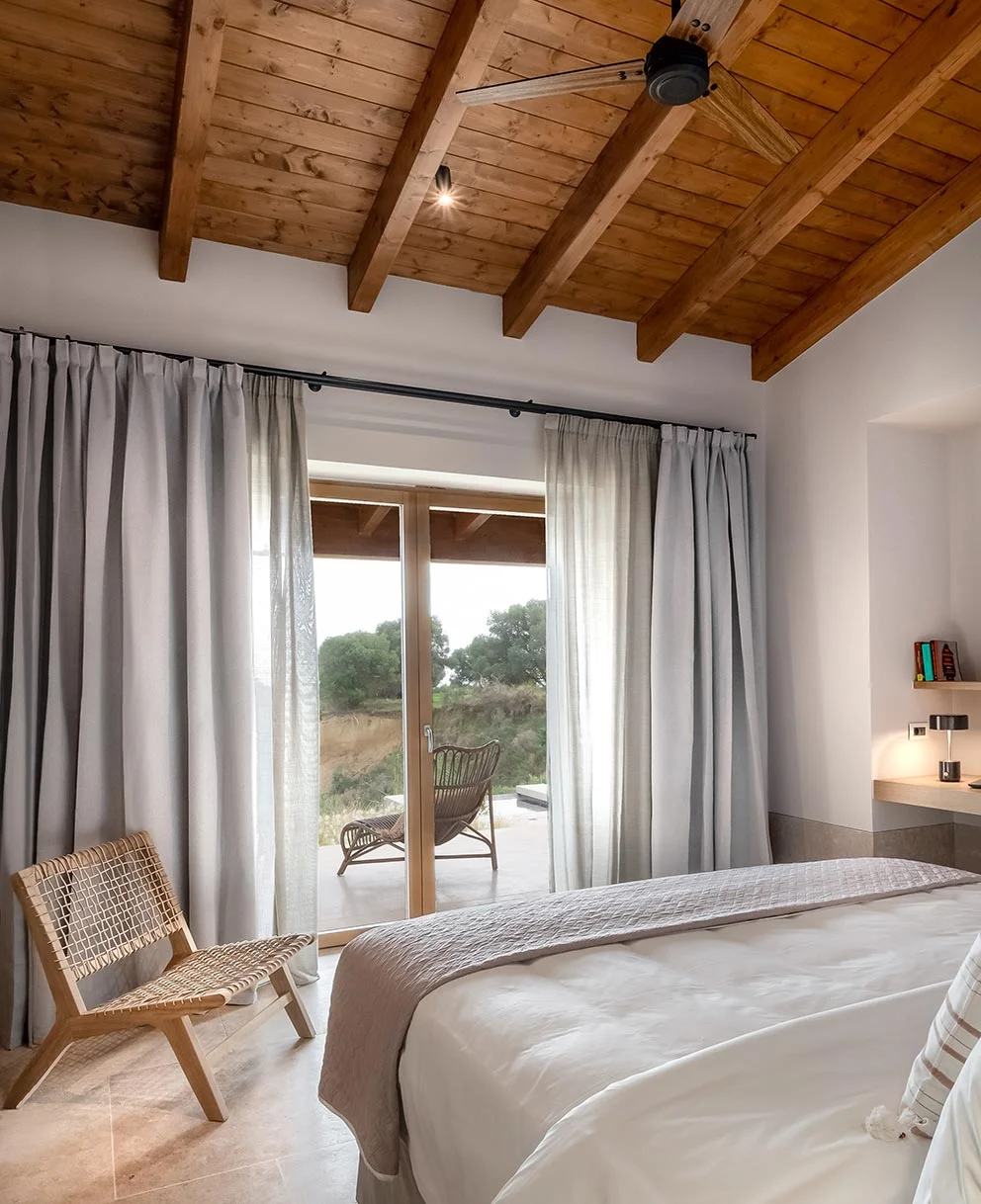 Bedroom with large windows, natural light, and rattan chair facing garden at Eliamos resort in Kefalonia.