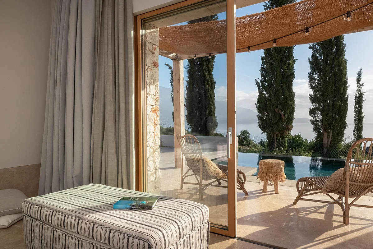 Glass doors open to sunlit private pool area with wooden lounge chairs at Eliamos in Kefalonia.